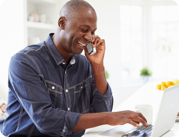 A man sitting at his kitchen table talking on the phone and using his laptop to browse the Internet