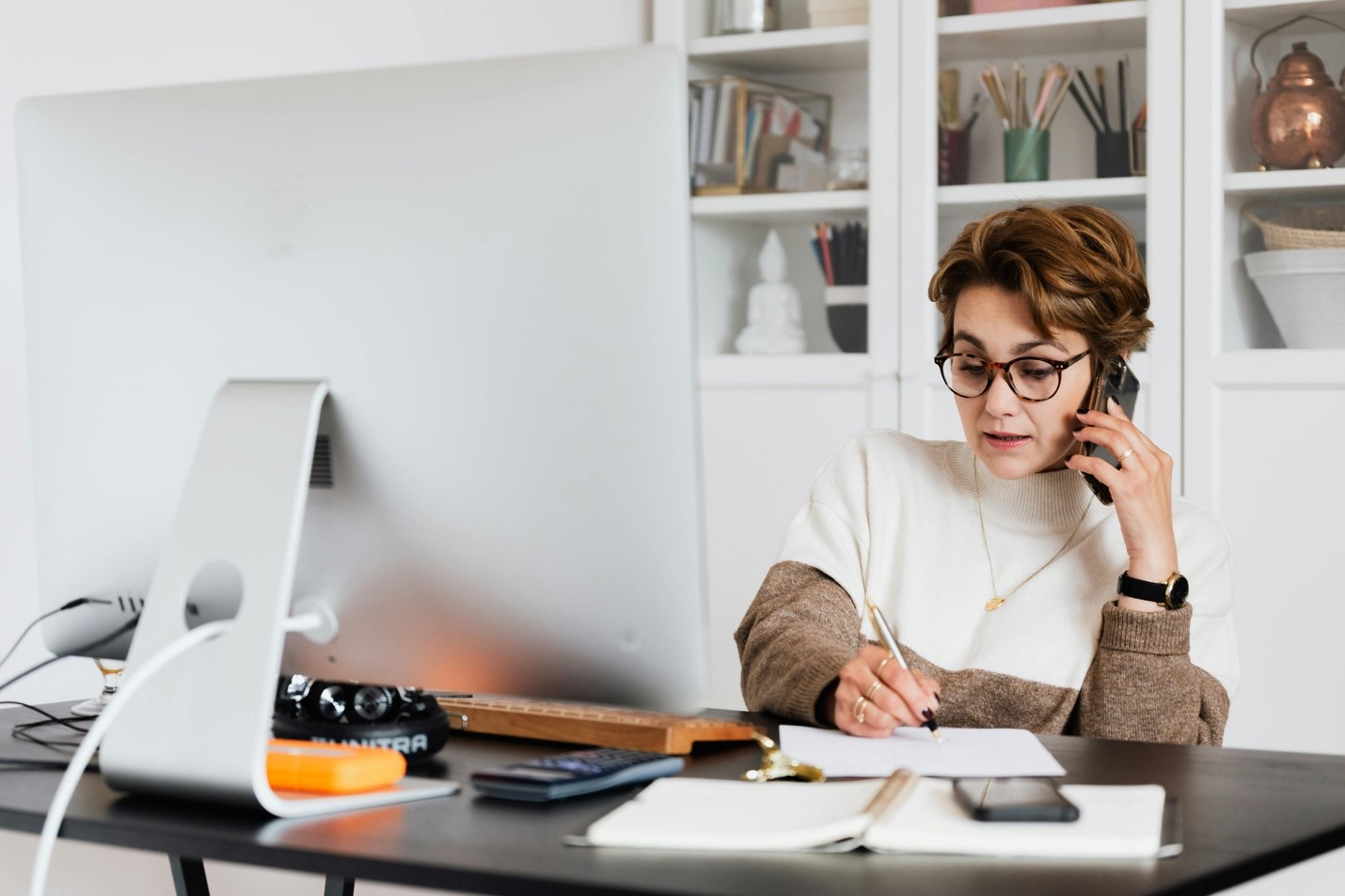 Woman working remotely, taking a phone call at home office computer.